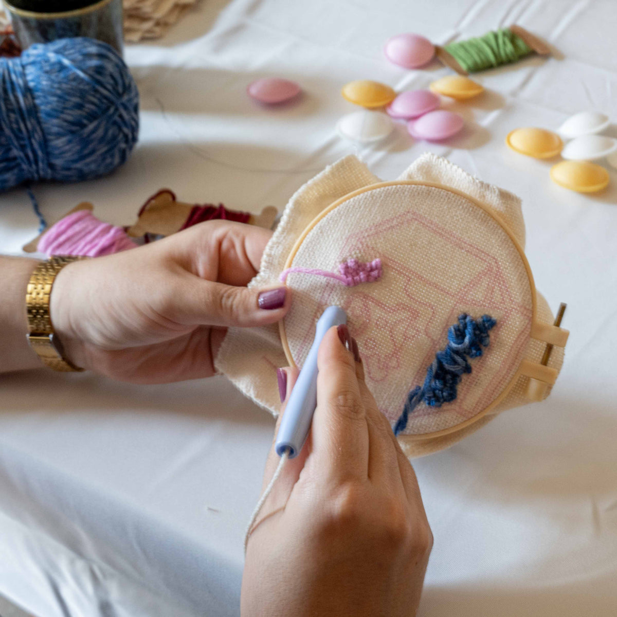 A close-up of hands using a crochet hook to stitch a pink floral design onto a beige fabric hoop, with blue and pink thread partially completed. The background shows a white tablecloth with colorful balls of yarn and small decorative items.
