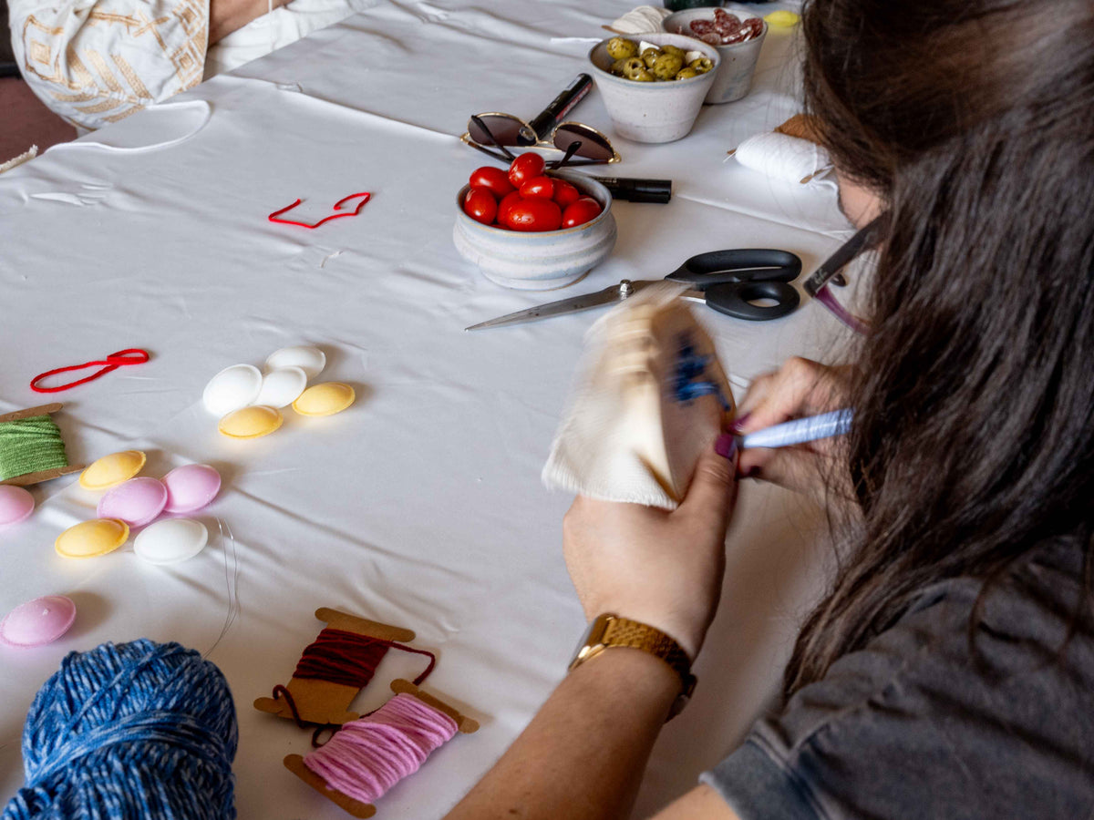 A person with long dark hair sits at a white table, holding a small beige fabric pouch and writing on it with a punch needle pen. The table is covered with craft supplies, including colorful yarn balls, spools of thread, scissors, sunglasses, and bowls of small decorative items.