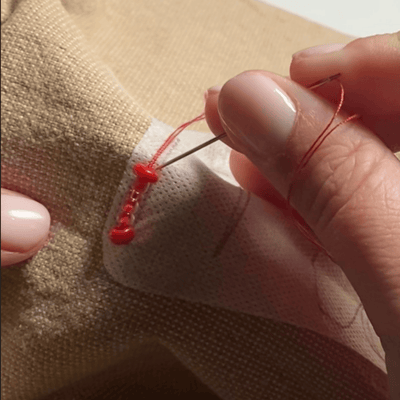 Hand sewing a red beads onto beige fabric with a beads embroidery needle.